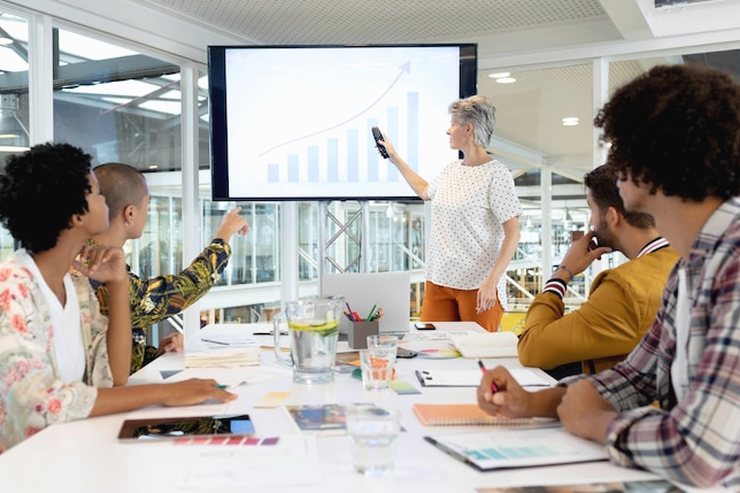A woman presenting a rising chart on a wall screen to a team seated around a long meeting table. Illustrative stock photo.