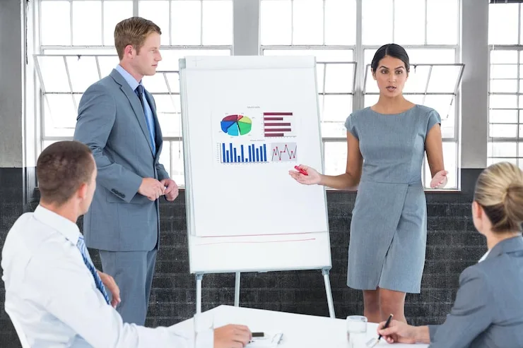 A man and a woman beside a flipchart showing pie and bar charts, with colleagues seated in front of them. Illustrative stock photo.