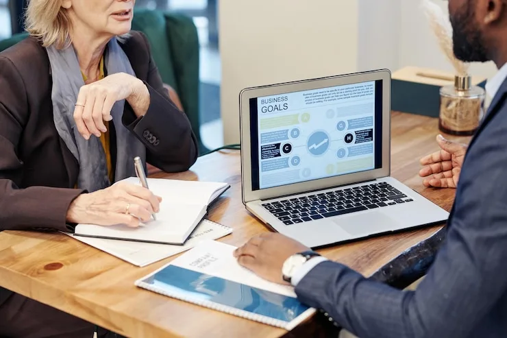 A woman taking notes in a notebook while two colleagues talk over a laptop on a wooden table. Illustrative stock photo.