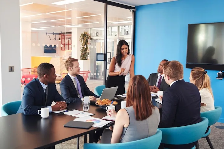 A woman presenting to colleagues seated around a conference table in a bright office with a blue wall. Illustrative stock photo.