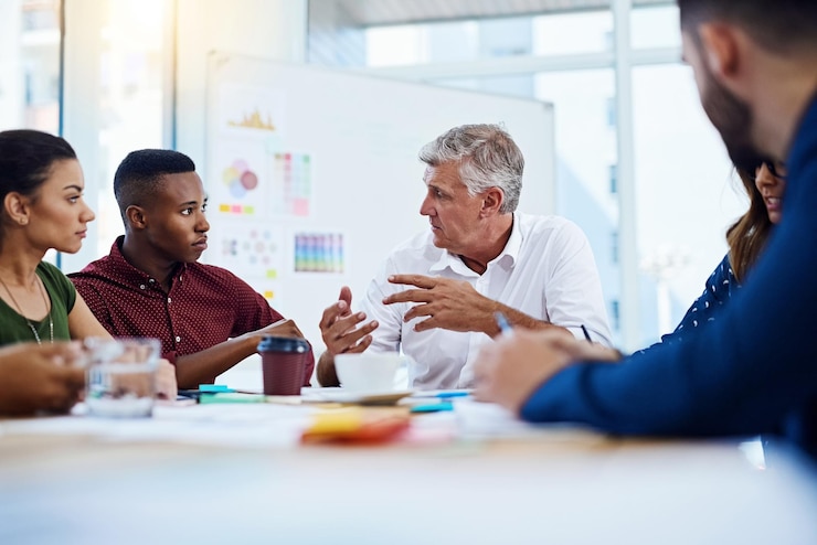 Six people talking around a meeting table, an older man gesturing while the others listen, with a whiteboard of notes behind them. Illustrative stock photo.